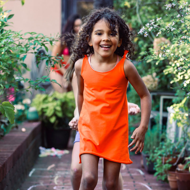 Girls in an orange cotton play dress running outdoors with plants in the background.