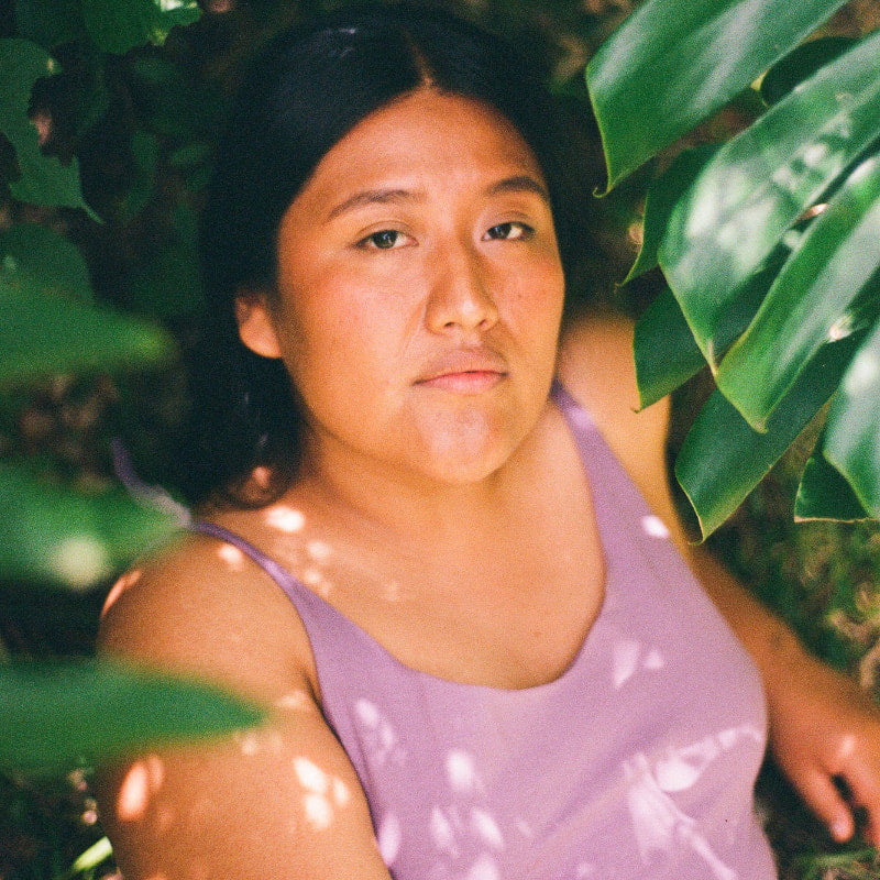 Woman wearing a cotton purple one piece standing among green leaves.