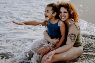 Woman and child sitting on a beach with bubbles on cotton one piece bathing suits, smiling.