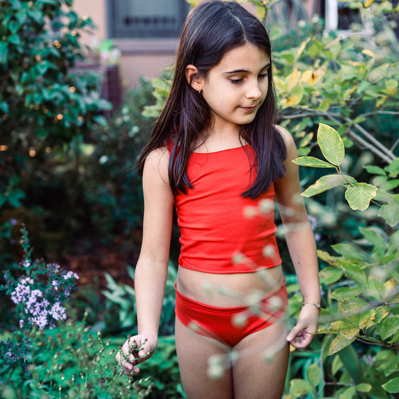 Young girl in a red cotton swimsuit standing among green plants.