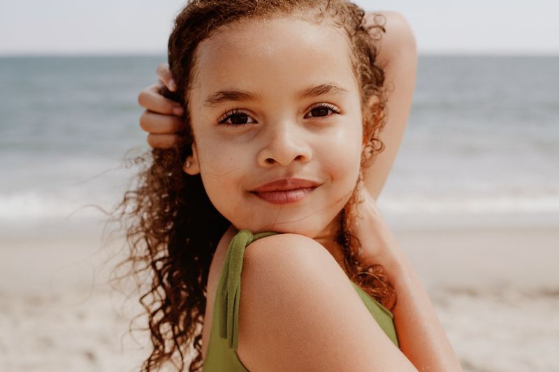Young girl with curly hair sitting on a beach, smiling at the camera.