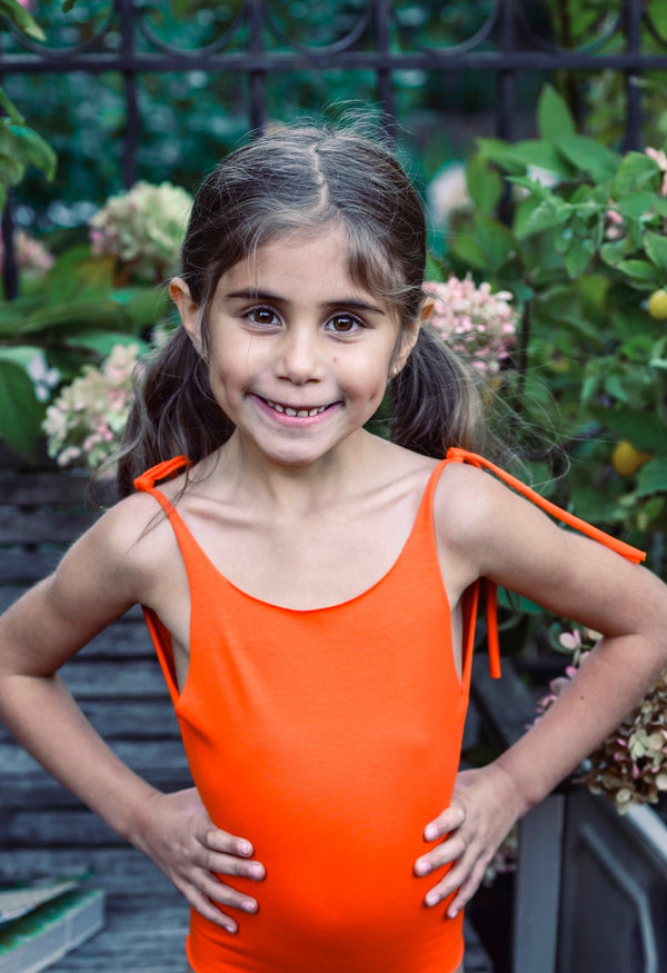 Young girl in an orange cotton one piece standing outdoors with greenery in the background